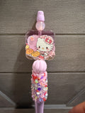 A pink Hello Kitty decorative item with a clear square charm featuring Hello Kitty and pastries, attached to a pink handle covered in colorful rhinestones, shown against a gray paneled background.