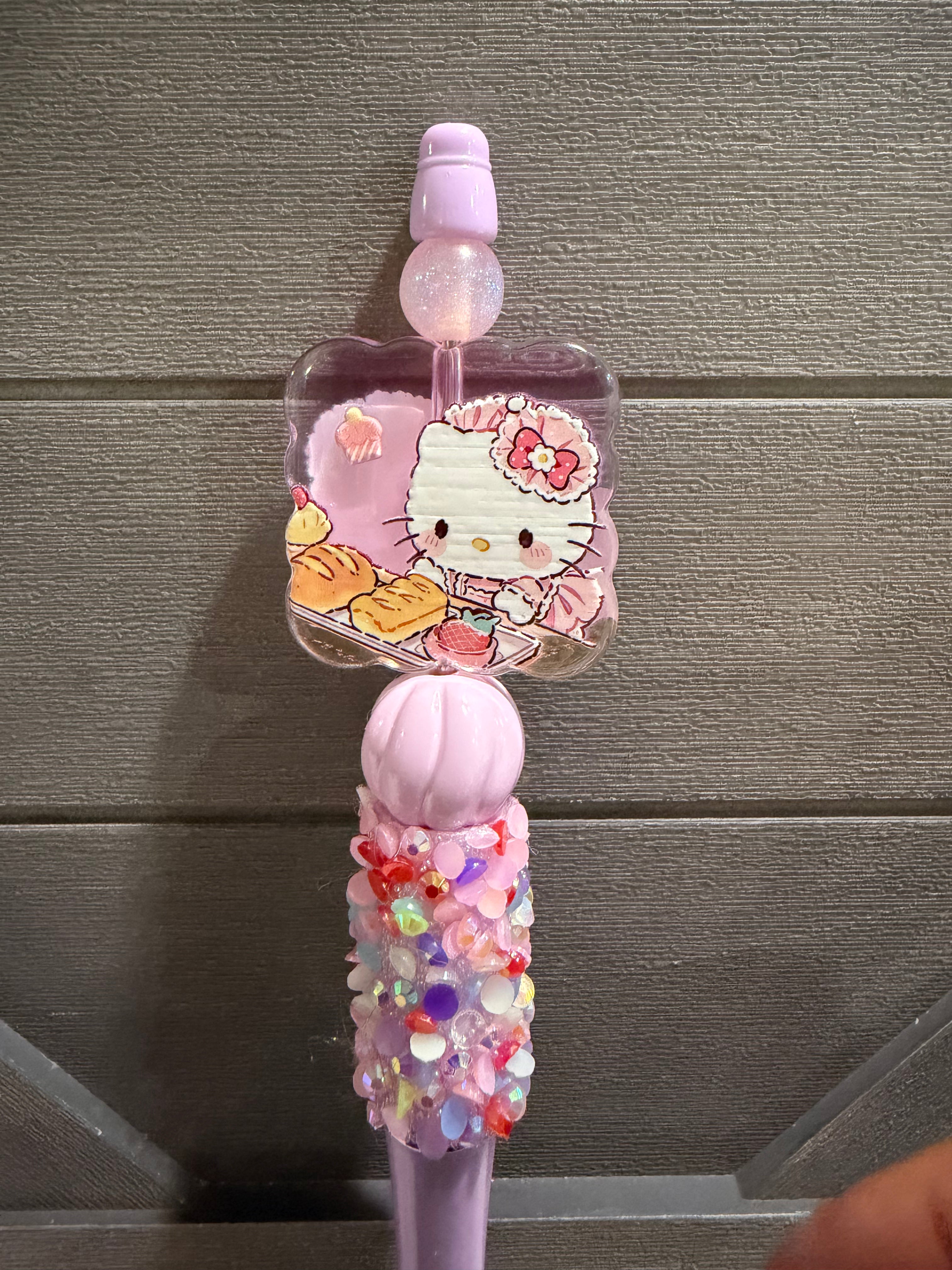 A pink Hello Kitty decorative item with a clear square charm featuring Hello Kitty and pastries, attached to a pink handle covered in colorful rhinestones, shown against a gray paneled background.