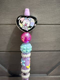 A decorative pastel purple pen or pencil with a heart-shaped cartoon bear topper, a pink translucent bead, a light blue faceted bead, and small flower and heart embellishments, shown against a gray wood background.