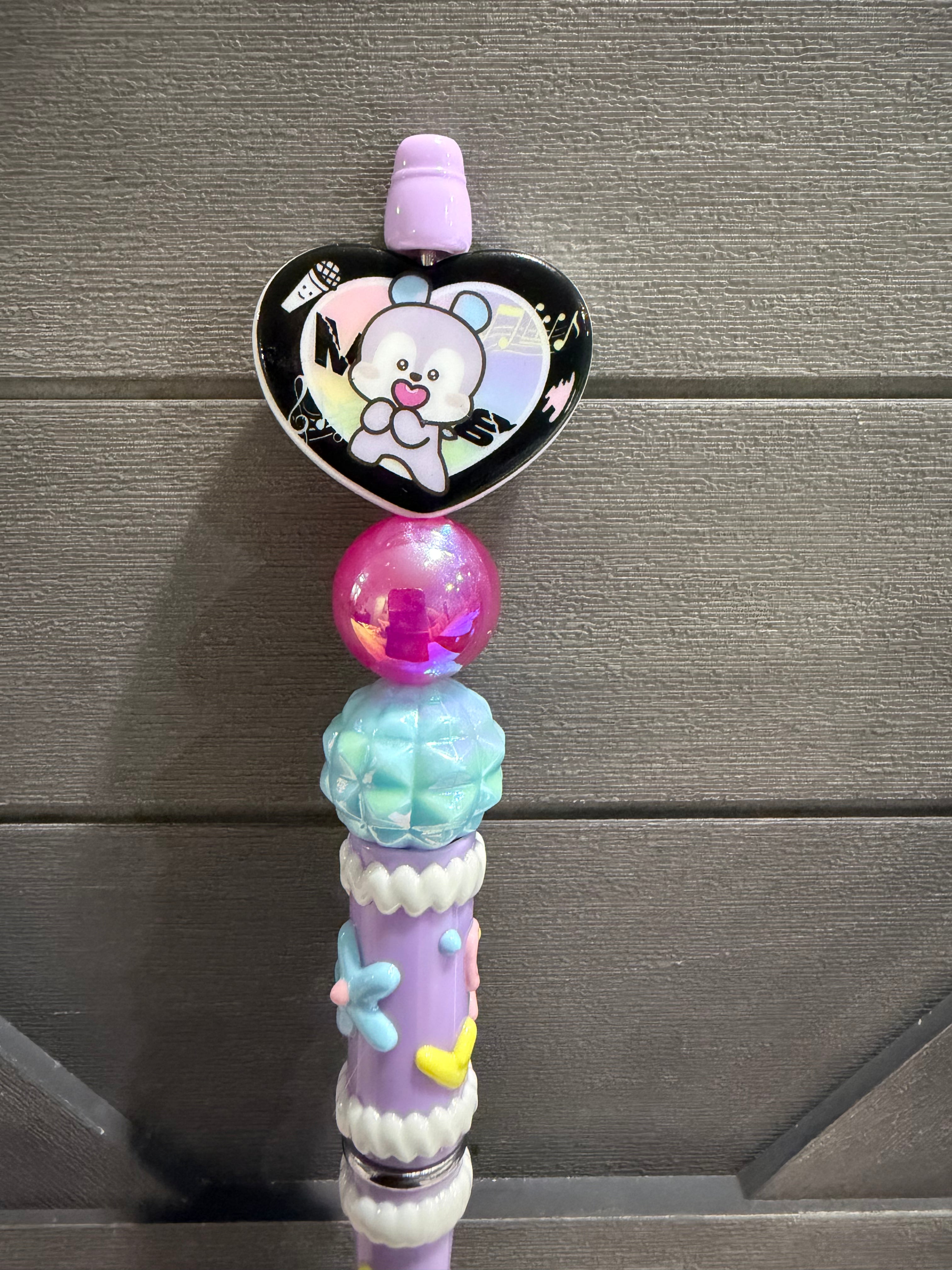 A decorative pastel purple pen or pencil with a heart-shaped cartoon bear topper, a pink translucent bead, a light blue faceted bead, and small flower and heart embellishments, shown against a gray wood background.