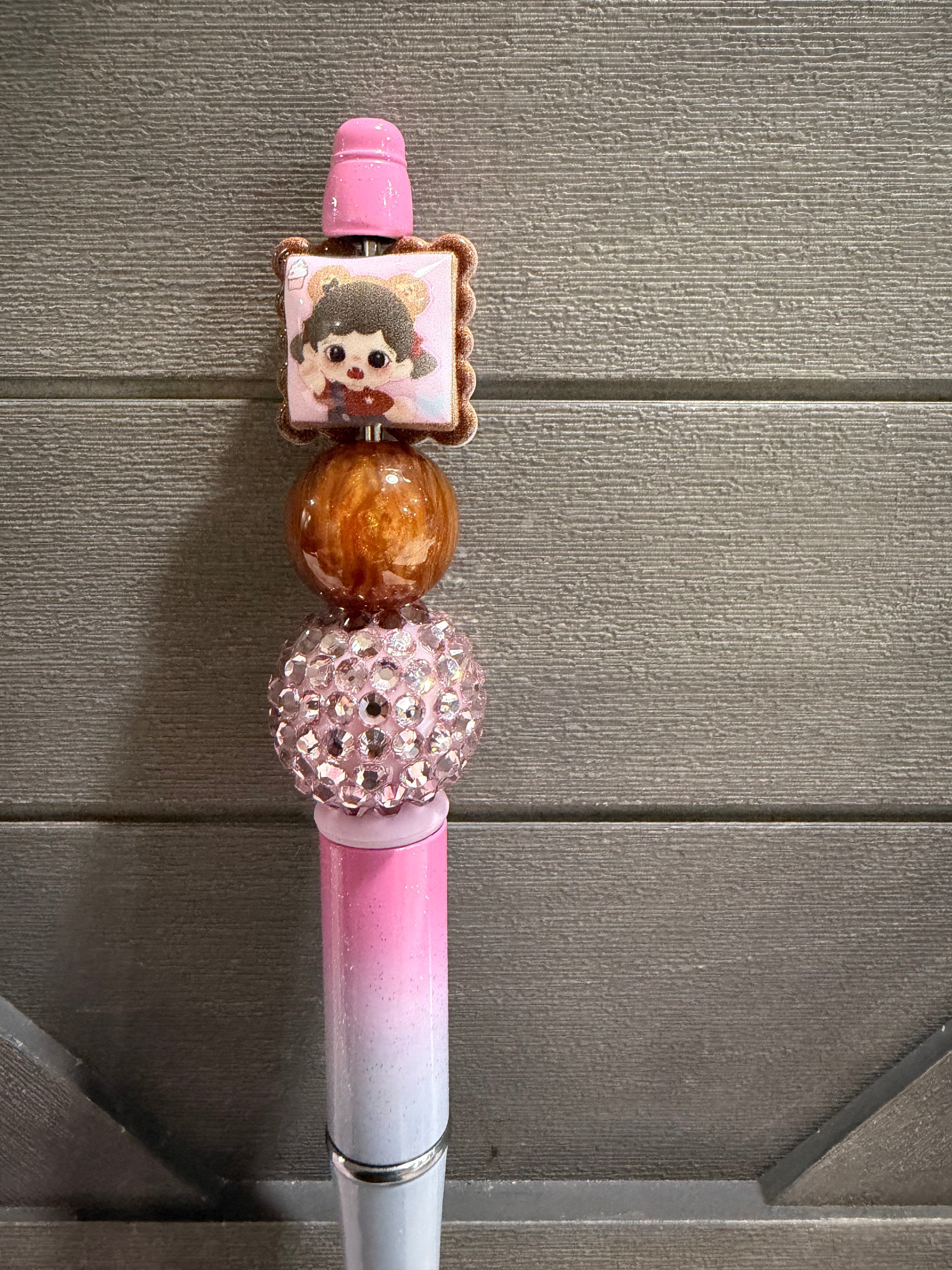 A decorative pink and white pen with a glossy pink cap, a square charm featuring a cartoon character, a brown marbled bead, a pink rhinestone ball accent, and a silver metal band near the bottom, set against a gray wood-paneled background.