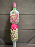 A close-up of a decorative pink and green accessory with a cartoon character printed on a rectangular piece, a bright pink bead, and a floral knotted section, set against a gray wood background.