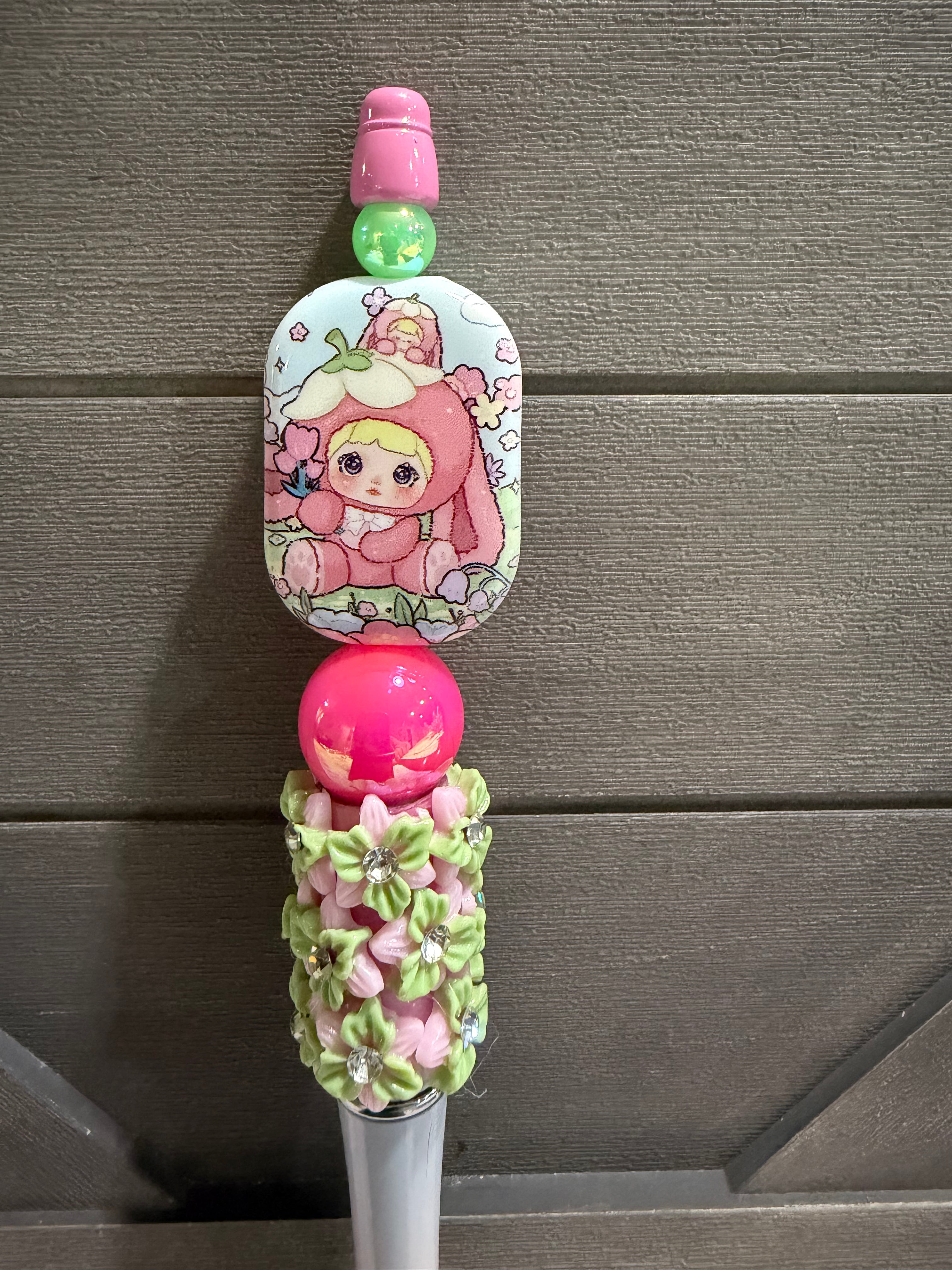 A close-up of a decorative pink and green accessory with a cartoon character printed on a rectangular piece, a bright pink bead, and a floral knotted section, set against a gray wood background.