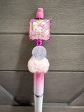 A pink and white Hello Kitty themed pen or stick-style accessory with a translucent square charm featuring Hello Kitty artwork, a fluffy pink pom-pom, and a metallic pink grip, shown against a gray paneled background.