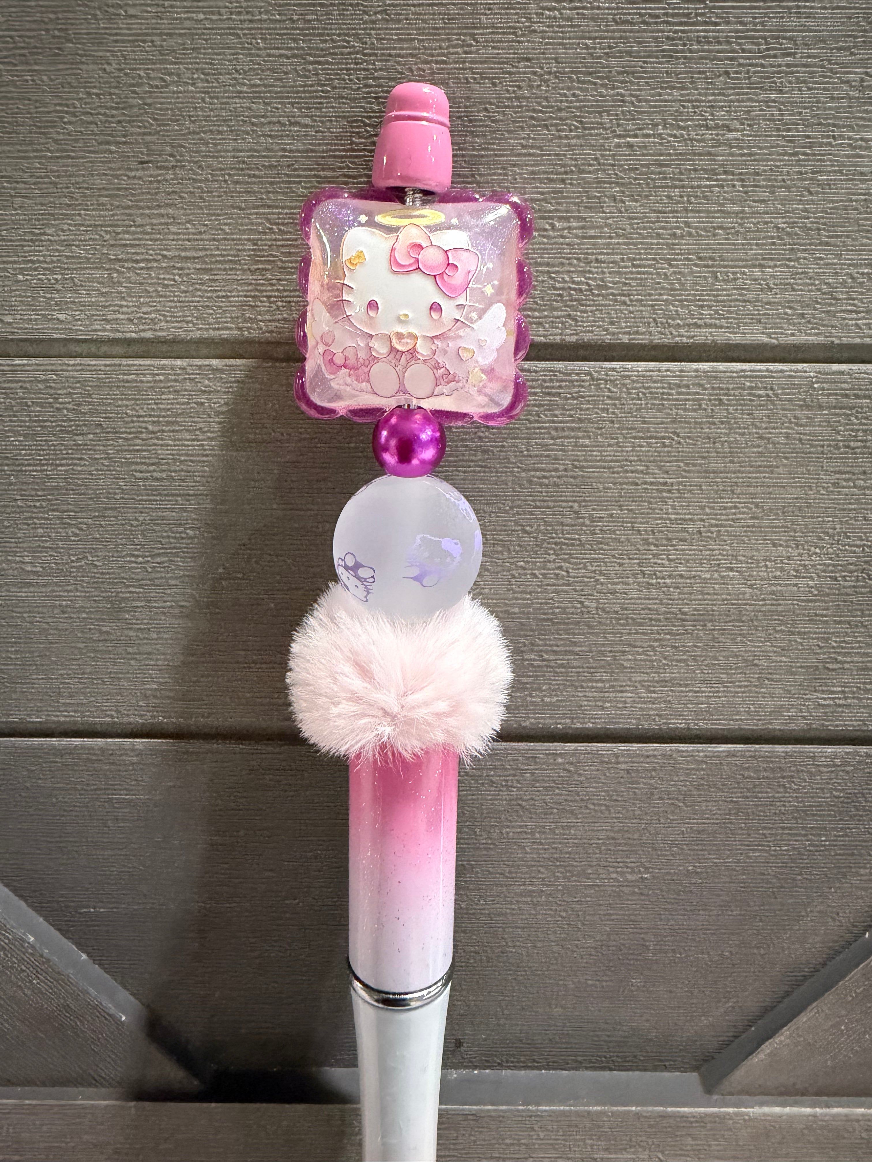 A pink and white Hello Kitty themed pen or stick-style accessory with a translucent square charm featuring Hello Kitty artwork, a fluffy pink pom-pom, and a metallic pink grip, shown against a gray paneled background.