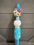 A close-up of a decorative blue plastic tool or accessory with a colorful flower-shaped charm featuring smiley faces, a white textured bead, a light blue iridescent bead, and a turquoise beaded section, set against a gray wood plank background.