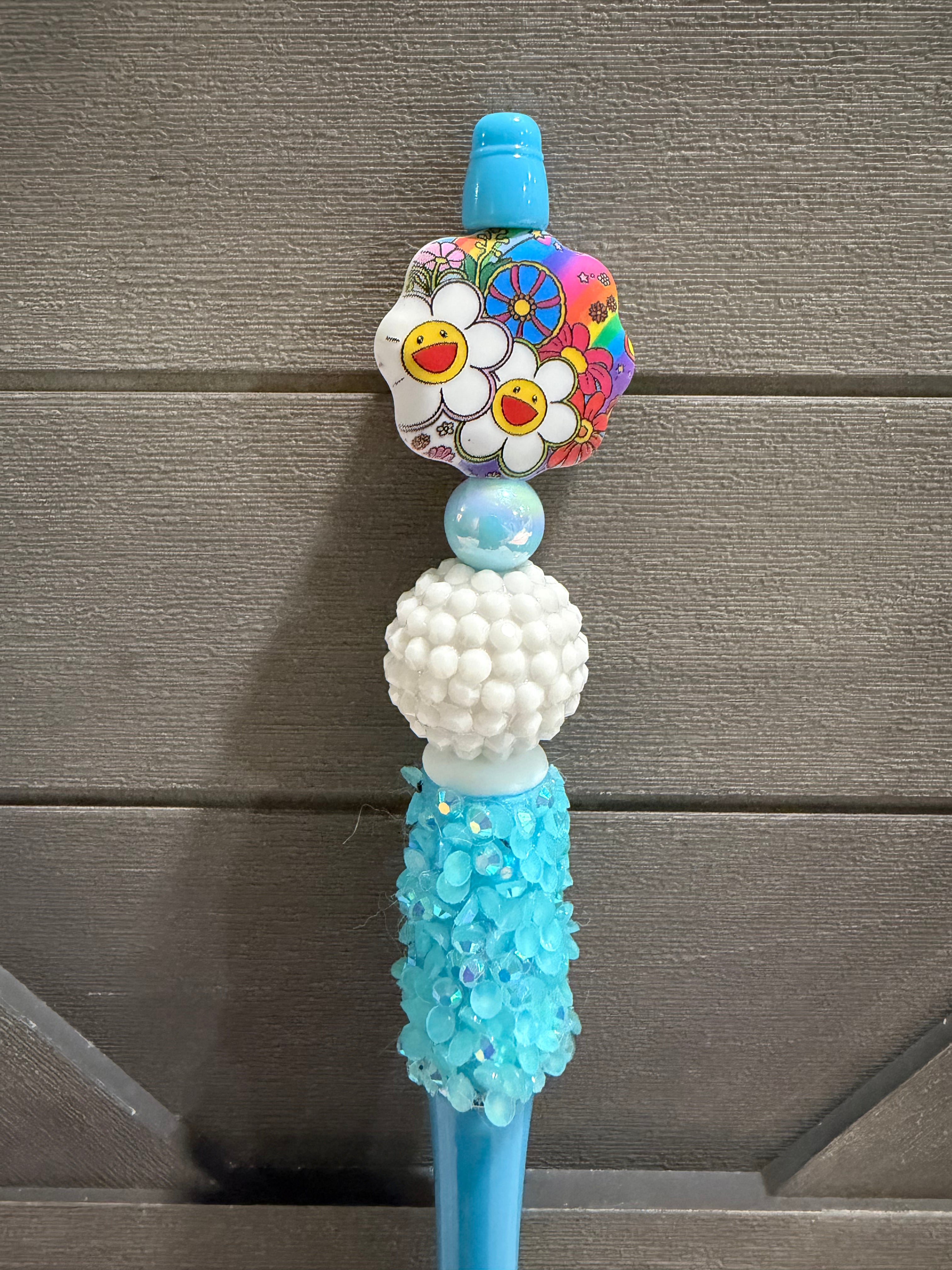 A close-up of a decorative blue plastic tool or accessory with a colorful flower-shaped charm featuring smiley faces, a white textured bead, a light blue iridescent bead, and a turquoise beaded section, set against a gray wood plank background.