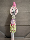 A decorative pink and white pen or stylus with a cartoon character charm, rhinestone ball, glittery purple sphere, and floral embellishments, shown against a gray wood plank background.