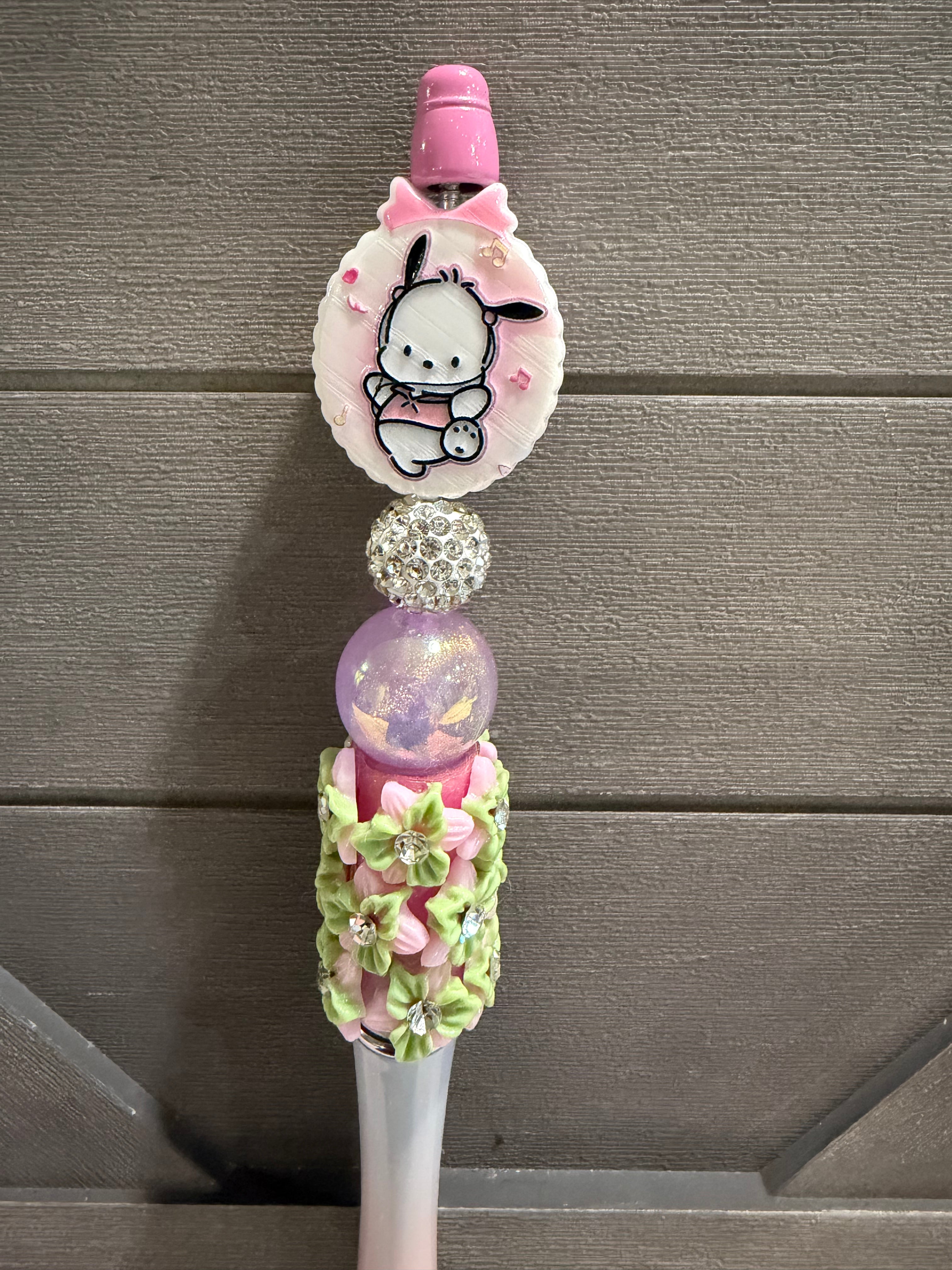A decorative pink and white pen or stylus with a cartoon character charm, rhinestone ball, glittery purple sphere, and floral embellishments, shown against a gray wood plank background.