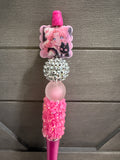 A pink Sailor Moon themed pen or pencil decorated with a character charm, a rhinestone ball, a translucent pink bead, and a pink flower-patterned grip, set against a dark gray wooden background.