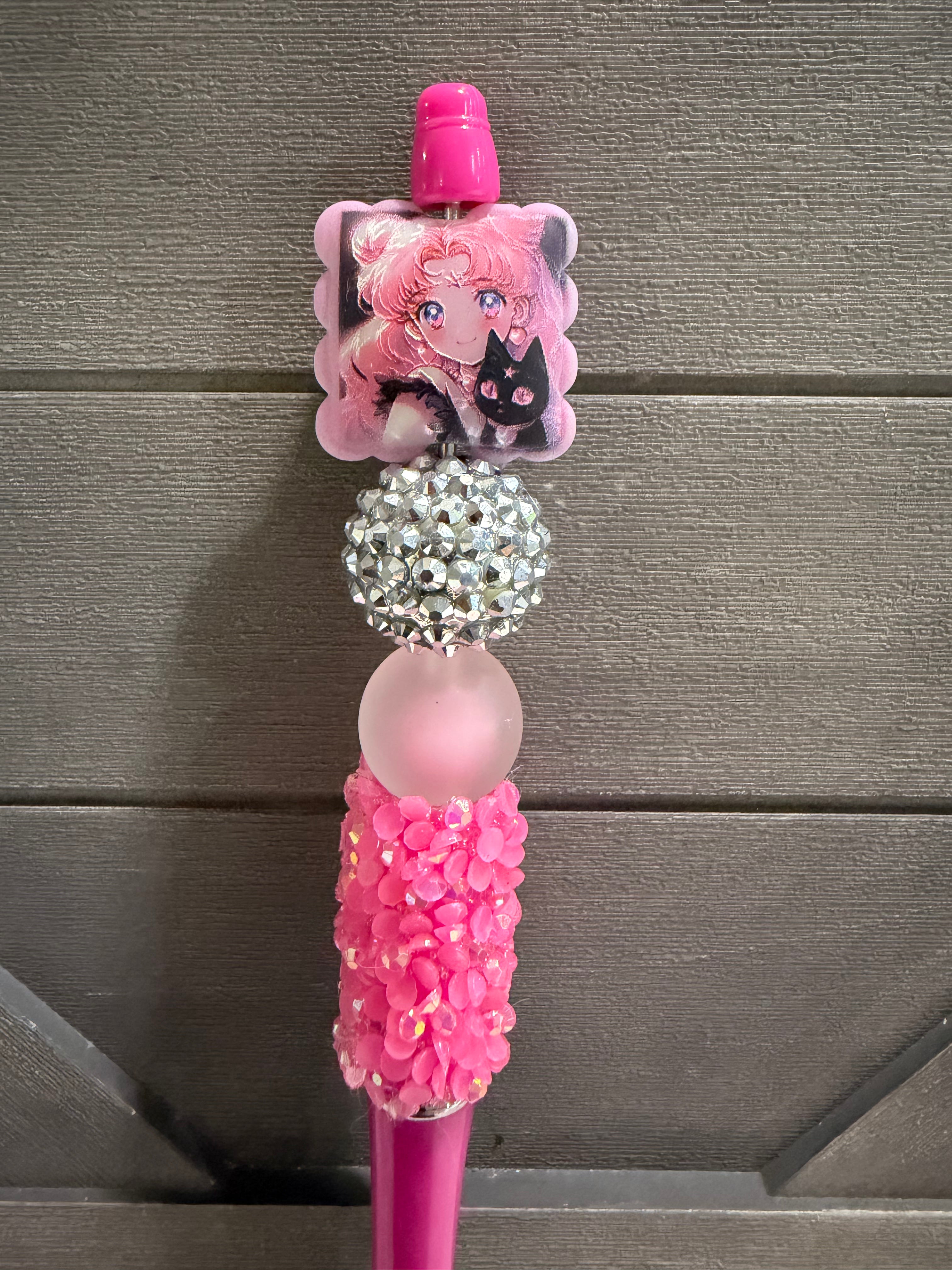 A pink Sailor Moon themed pen or pencil decorated with a character charm, a rhinestone ball, a translucent pink bead, and a pink flower-patterned grip, set against a dark gray wooden background.