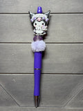 A purple ballpoint pen with a Kuromi character charm, a fluffy white pom-pom, and a glittery purple accent, displayed on a gray wooden background.