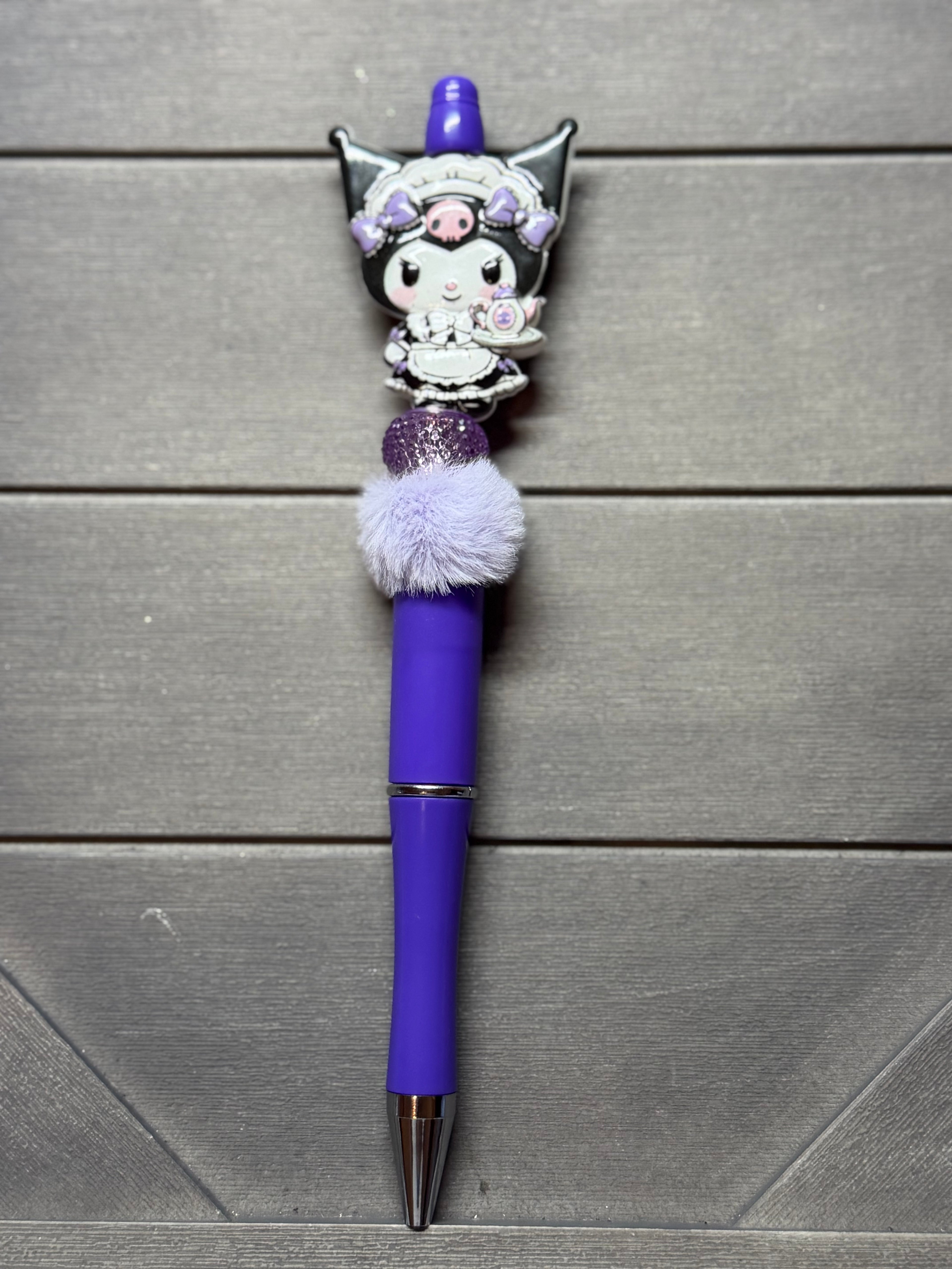 A purple ballpoint pen with a Kuromi character charm, a fluffy white pom-pom, and a glittery purple accent, displayed on a gray wooden background.