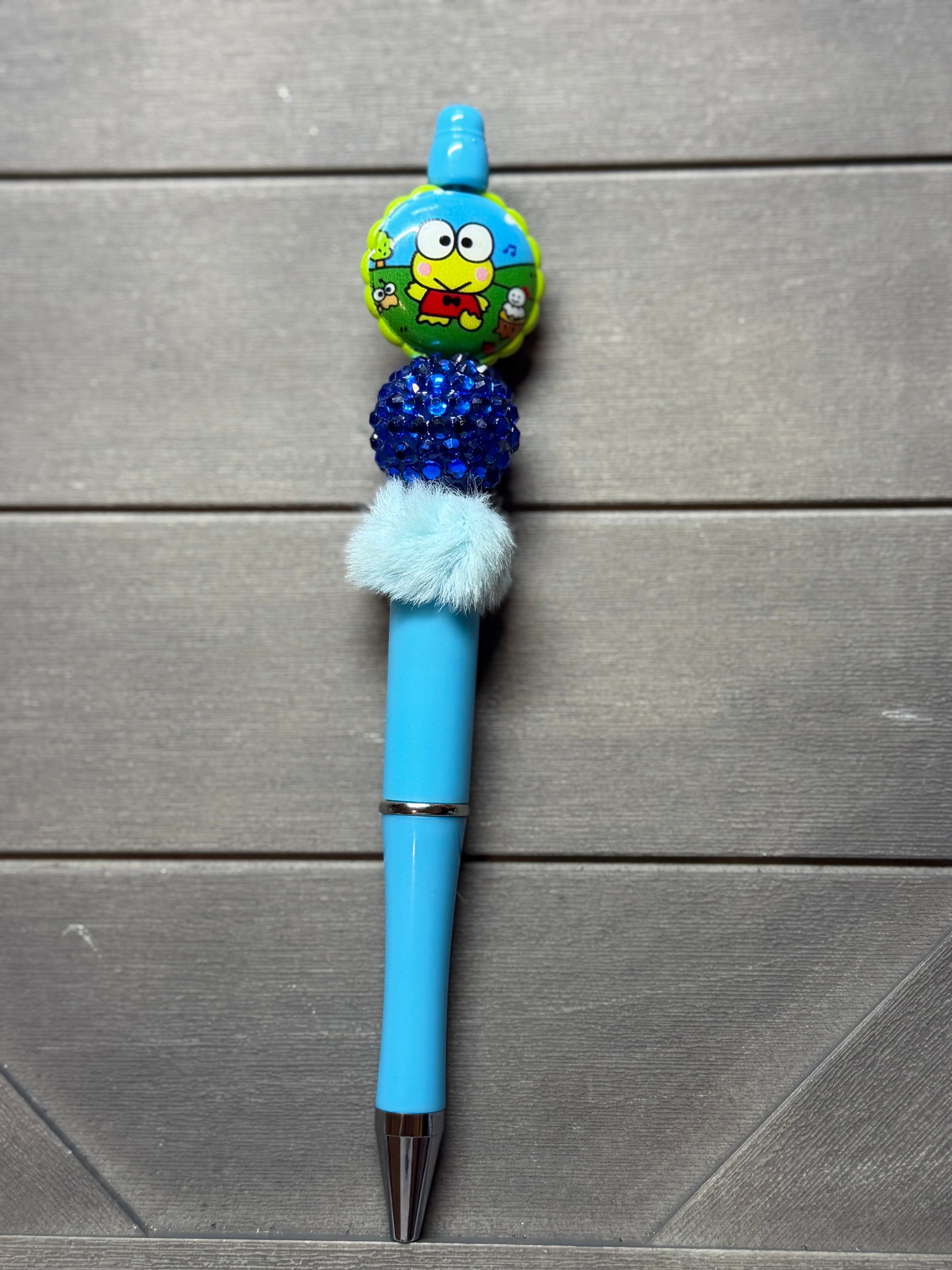 A light blue retractable pen with a decorative top featuring a cartoon character charm, a blue glittery pom-pom, a fluffy white accent, and a silver tip, displayed on a gray wood background.