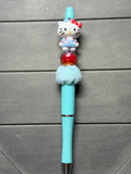 A light blue pen topped with a Hello Kitty figurine wearing a red bow, standing on a red circular detail, and a fluffy light blue pom-pom, set against a dark gray wood background.