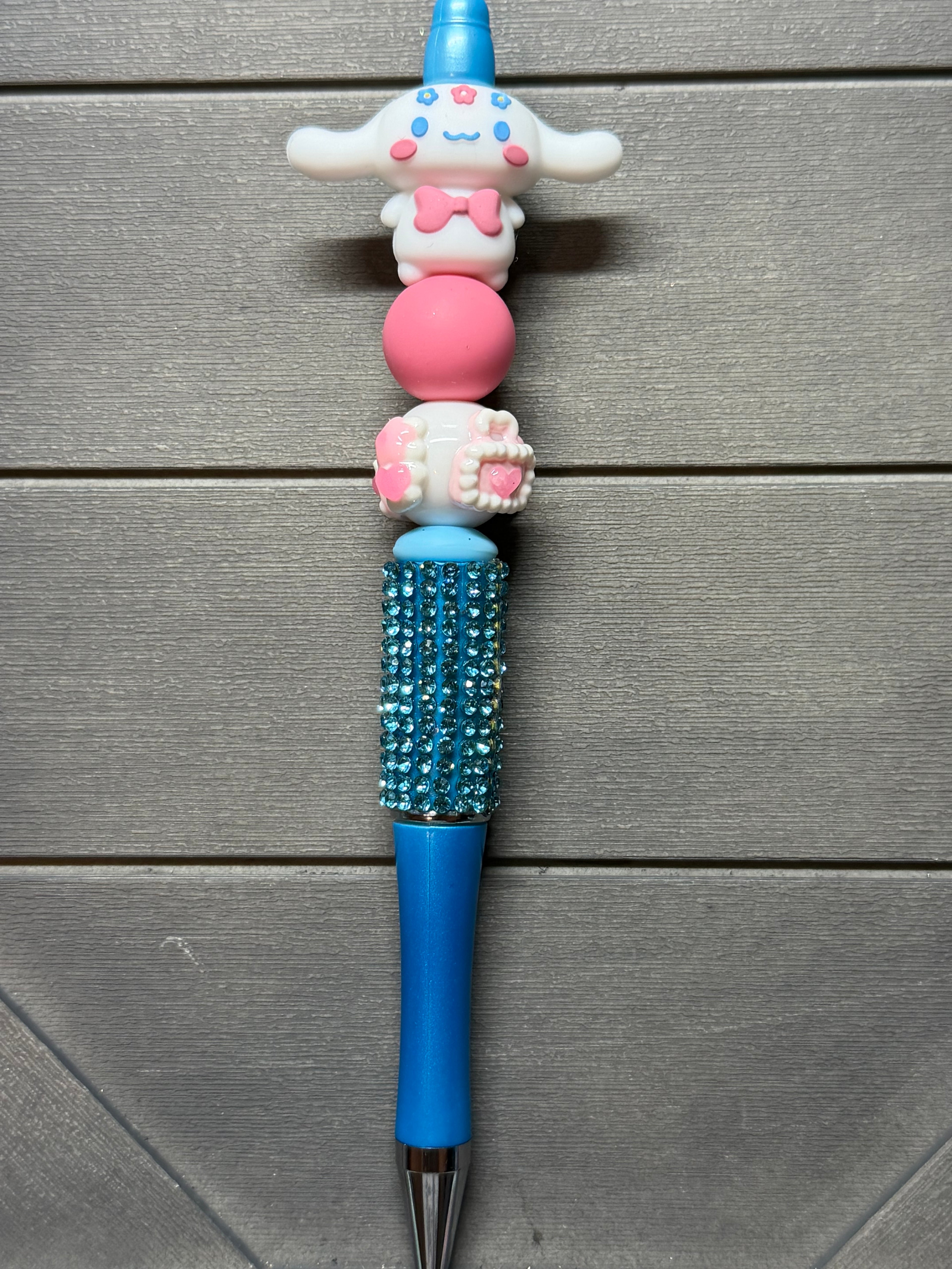 A blue and pink decorative pen with a cartoon character topper, a pink bead, a rhinestone-embellished blue grip section, and a silver tip, shown on a gray wood plank background.