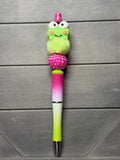 A novelty pen with a green cartoon frog character on top, a pink glittery pom-pom, a pink-to-green gradient barrel, and a lime green tip, displayed on a dark gray wood background.