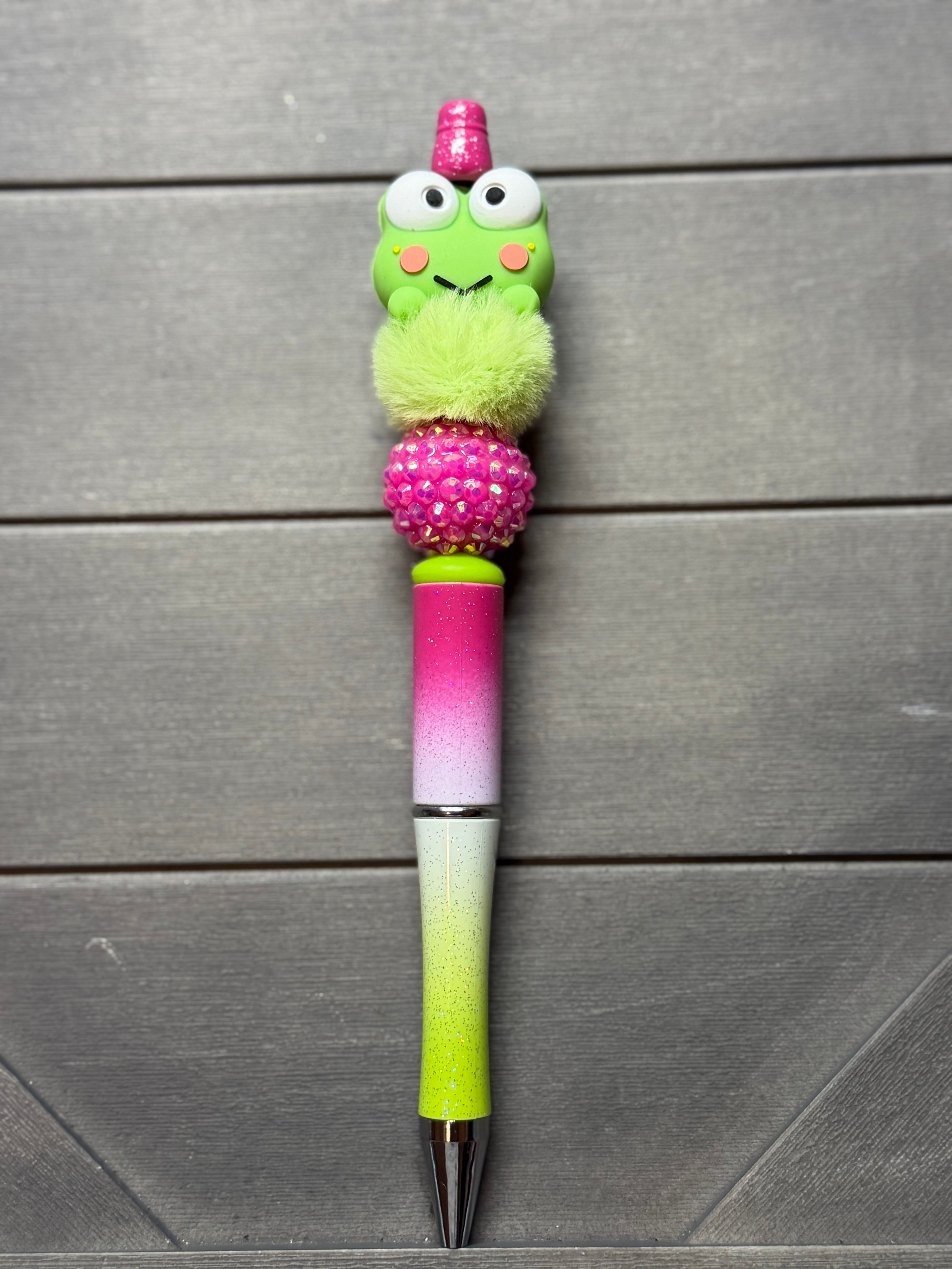 A novelty pen with a green cartoon frog character on top, a pink glittery pom-pom, a pink-to-green gradient barrel, and a lime green tip, displayed on a dark gray wood background.