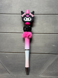 A decorative ballpoint pen with a Kuromi character topper, pink rhinestone details, a pink pom-pom, and a pink-to-white gradient barrel, displayed on a gray wood plank background.