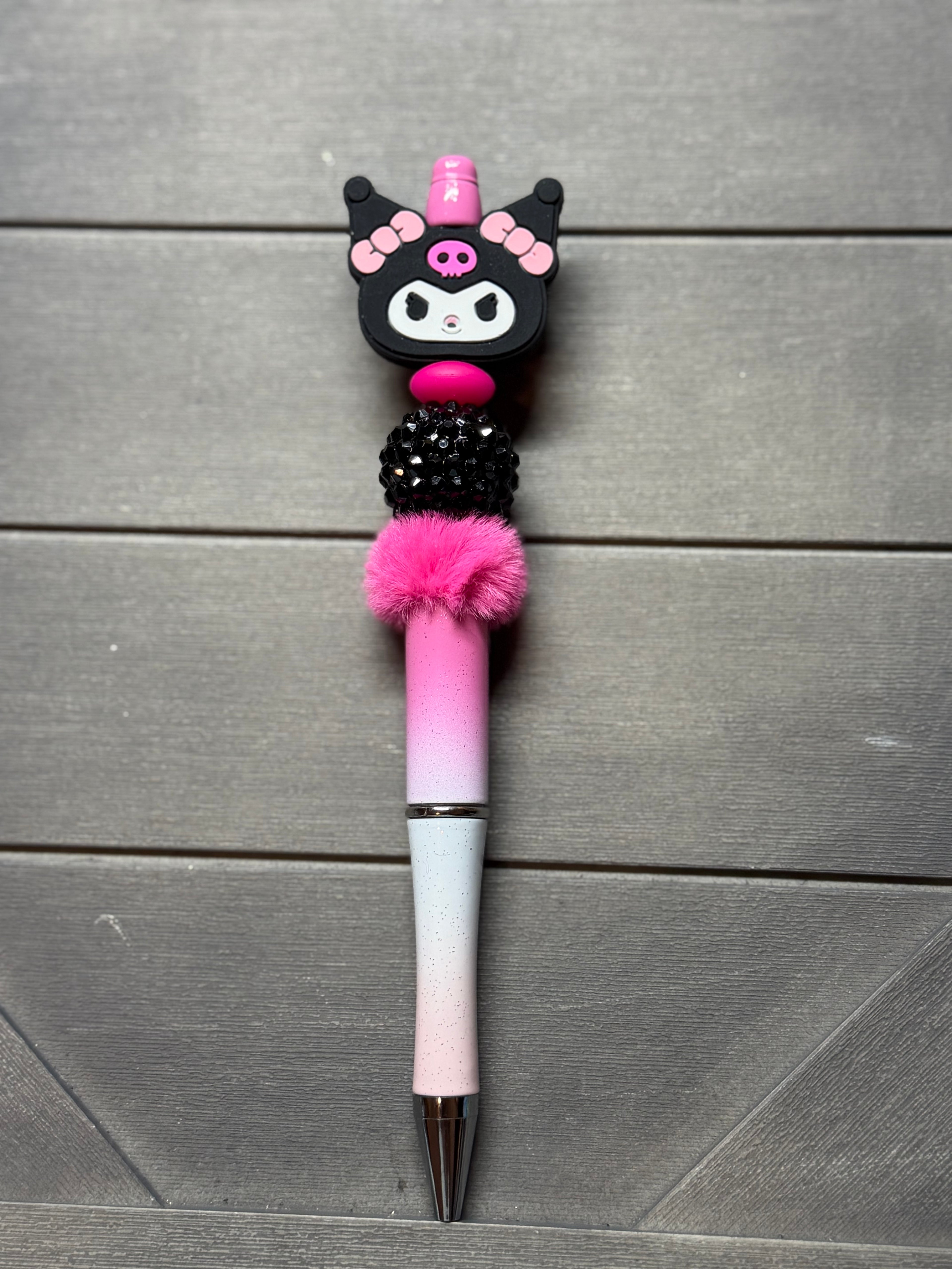 A decorative ballpoint pen with a Kuromi character topper, pink rhinestone details, a pink pom-pom, and a pink-to-white gradient barrel, displayed on a gray wood plank background.
