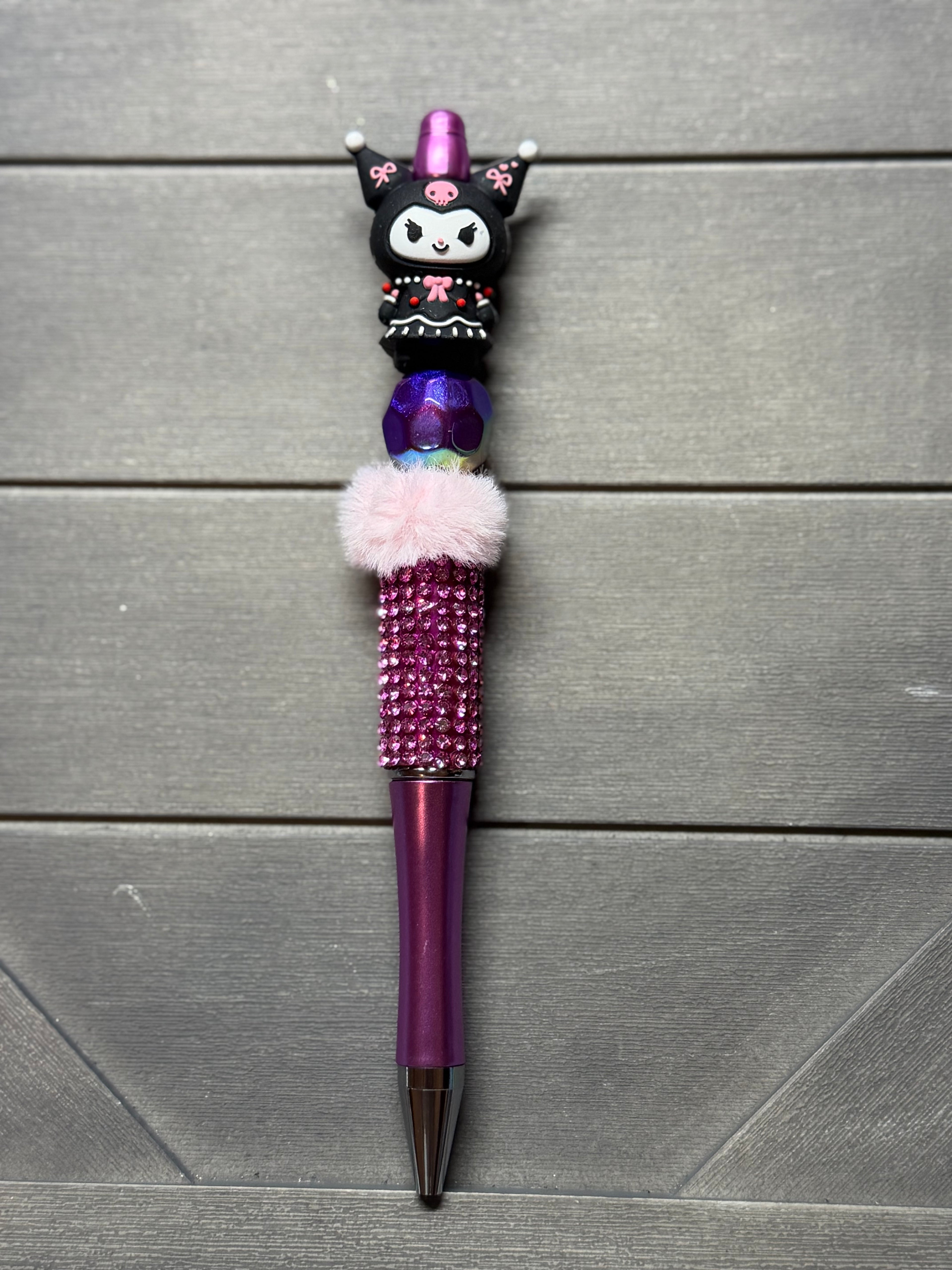 A purple and pink Kuromi-themed decorative pen with rhinestone detailing, a fluffy pink pom-pom, and a cartoon character topper, displayed on a gray wood plank background.